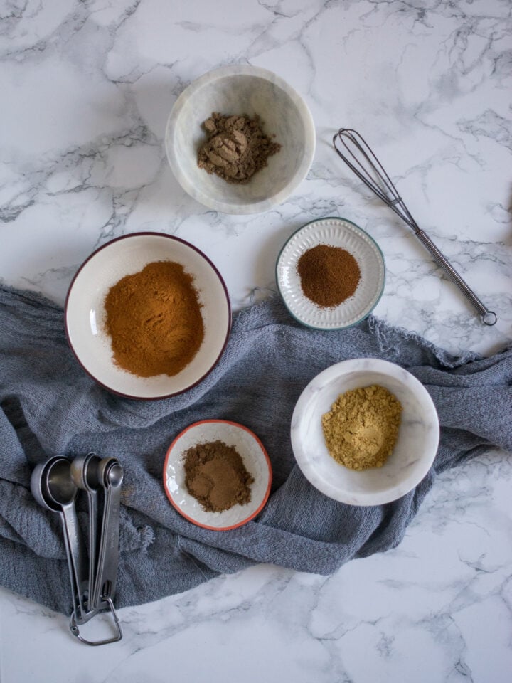 All the ingredients for homemade chai spice blend are in various glass bowls on a white marble backdrop with a gray towel.