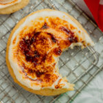 A creme brûlée sugar cookie with a bite taken out sits on a wire rack surrounded by small Christmas baubles and ribbon with other cookies.