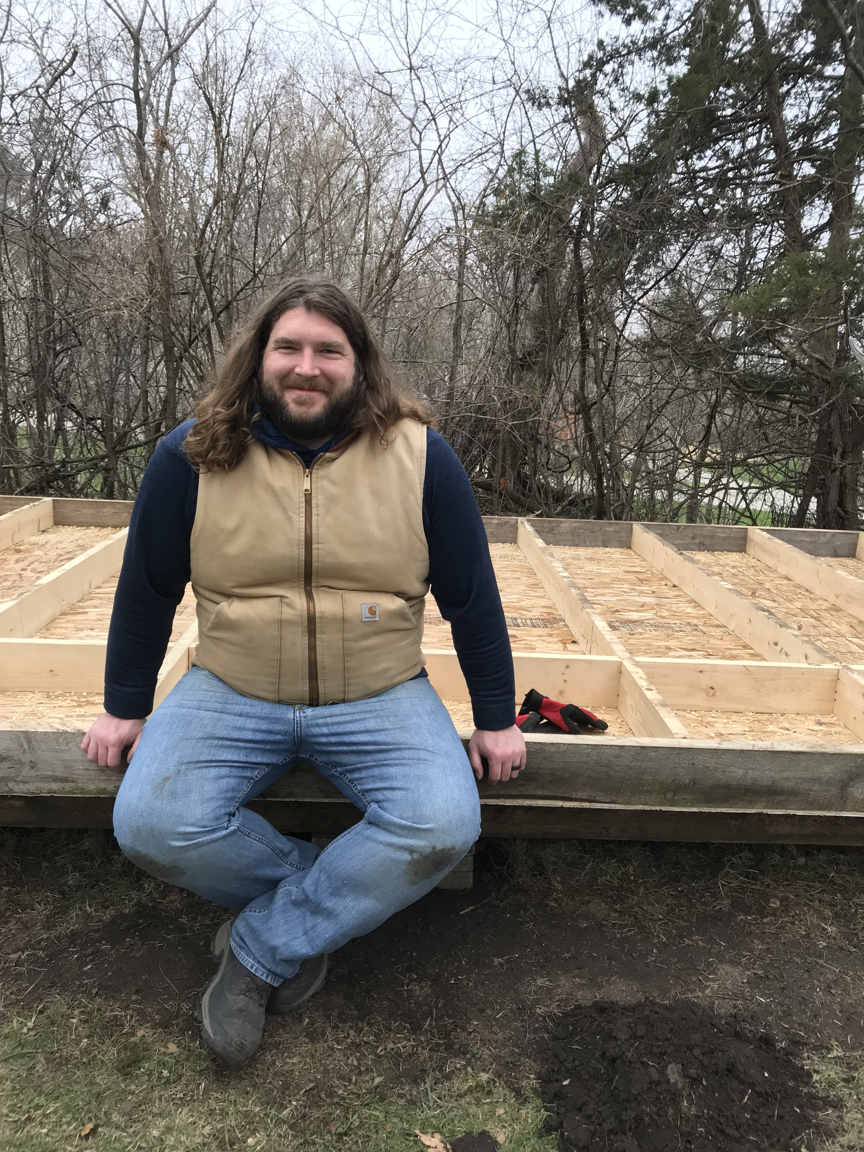 A bearded man with long hair wearing a tan Carhartt vest sits on the floor of what will be a garden shed. He is smiling.