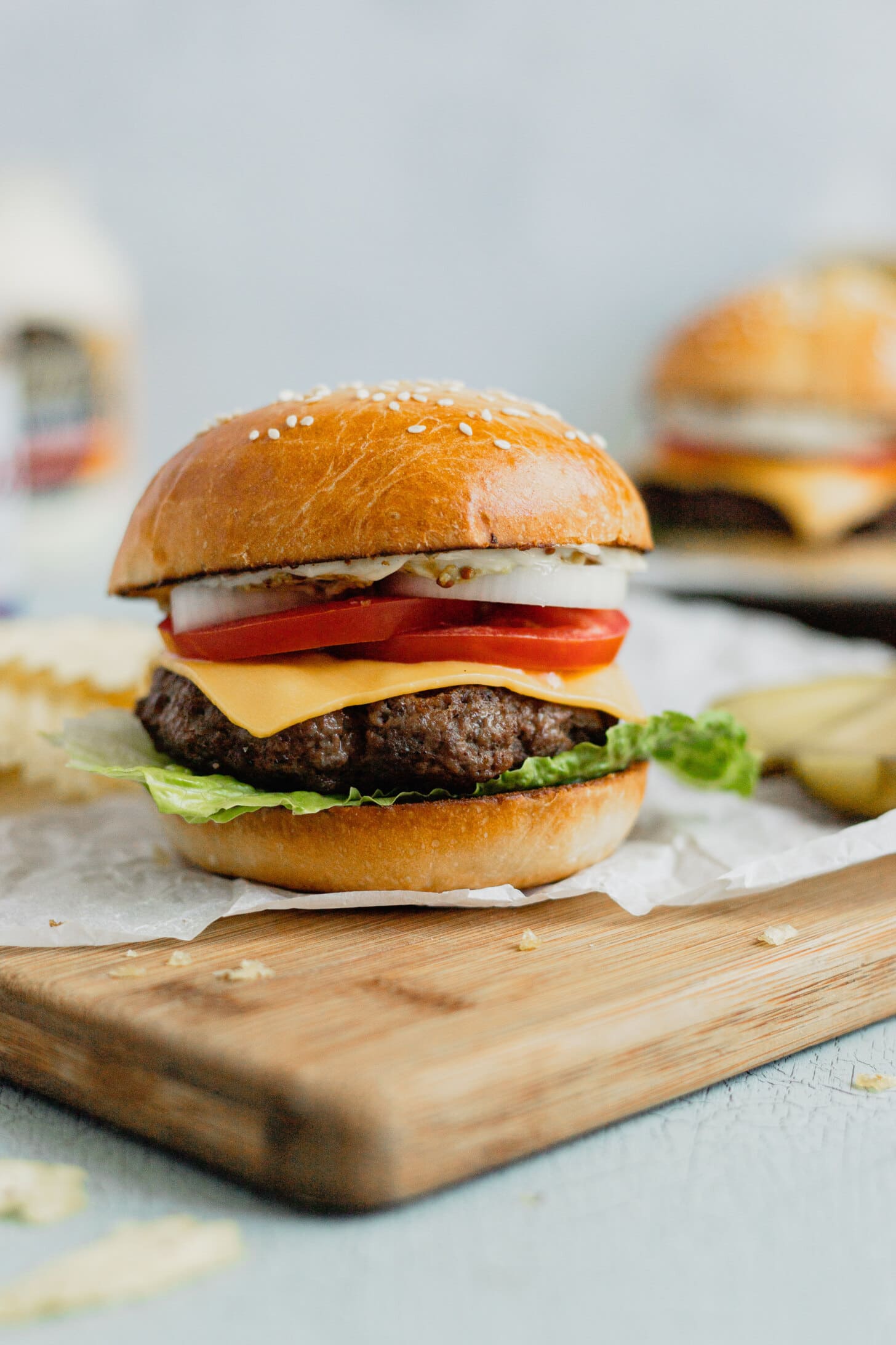  Una hamburguesa de venado molida se sienta en un bollo de brioche con lechuga, tomate, queso y cebolla. La hamburguesa está en un trozo de papel pergamino blanco sobre una tabla de cortar.