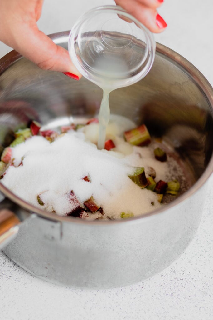 A hand pours lemon juice over sliced rhubarb and granulated sugar in a saucepan.