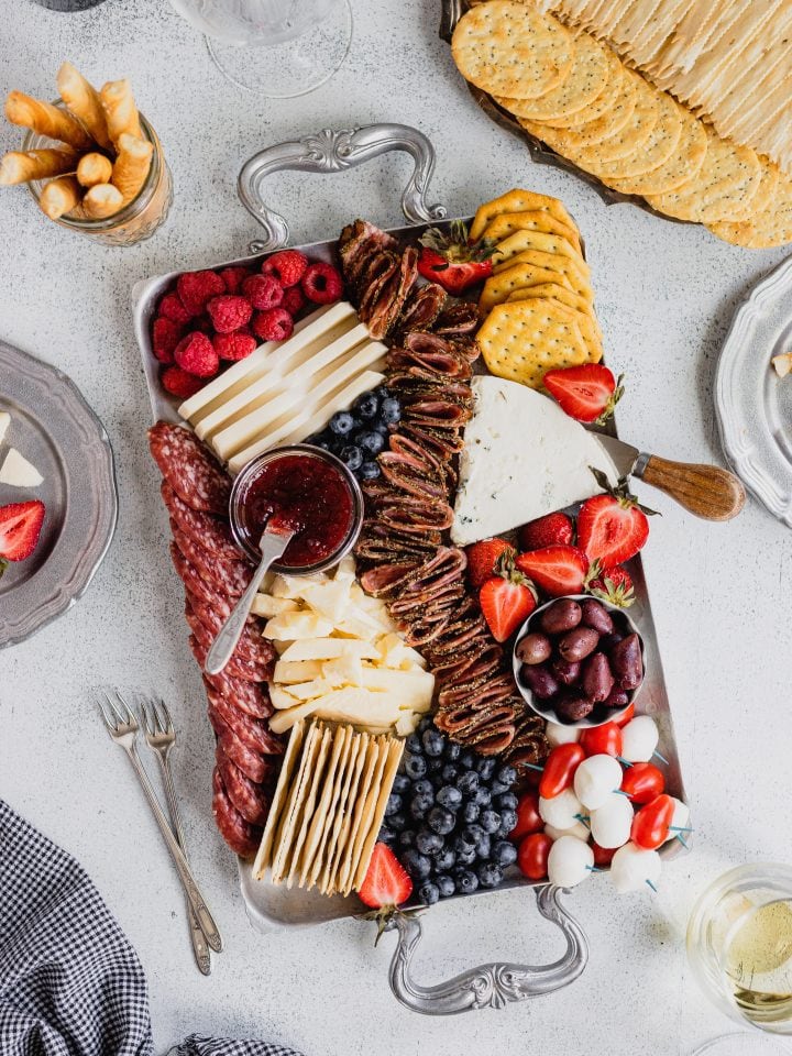 A silver tray is filled with red, white, and blue fruits, cheeses, and crackers to celebrate Fourth of July. There is a tray of crackers in the top right corner and wine glasses in the bottom right and top left.