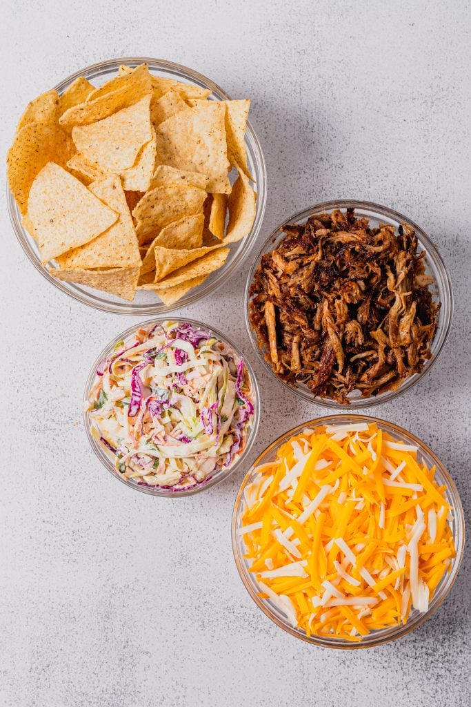 Four glass bowls hold the ingredients for homemade nachos. One bowl has tortilla chips, one has shredded pork, one has shredded cheese, and one has coleslaw.