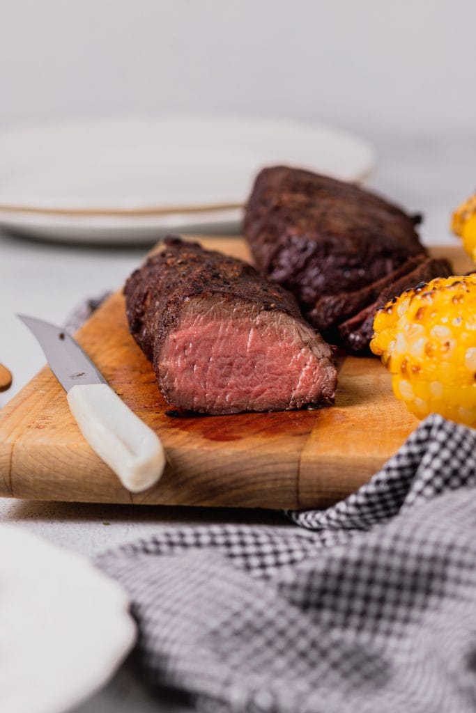 a straight on shot of a grilled venison backstrap that has been cut to expose the center. O veado repousa sobre uma tábua de corte de madeira com milho grelhado.