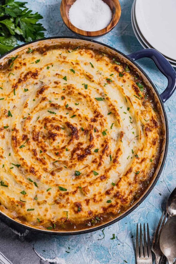 A shallow cast iron Dutch oven is filled with venison shepherd's pie. The top is sprinkled with parsley and the pan rests on a blue backdrop.