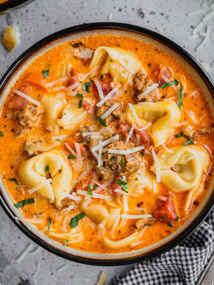 A bowl of sausage and tortellini soup is garnished with shaved parmesan.