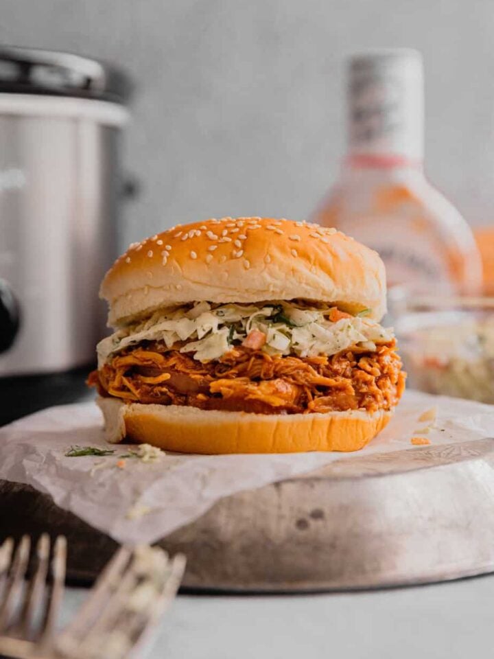A shredded bbq chicken sandwich sits on a metal plate in front of a crockpot.