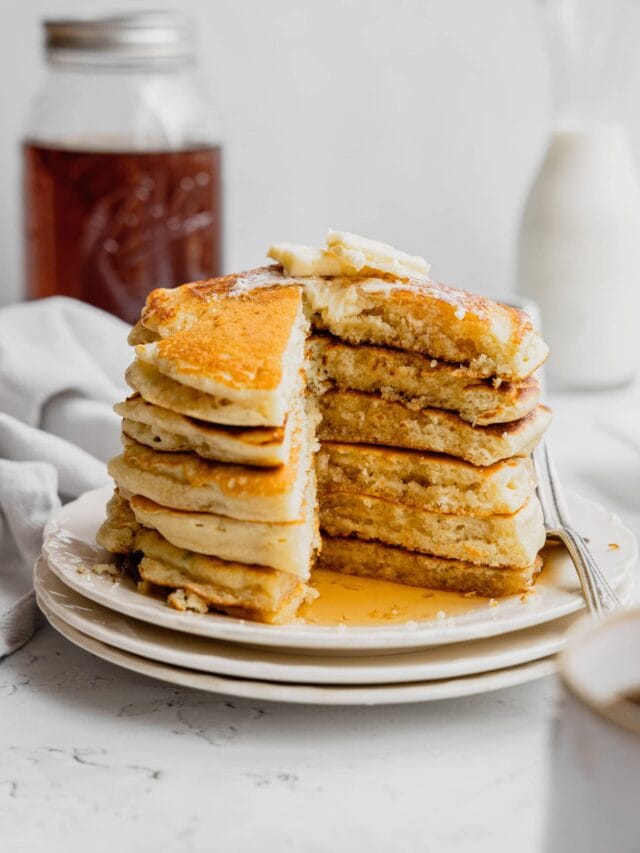 A stack of buttermilk pancakes on a white plate with a piece cut out to show the tender interior.