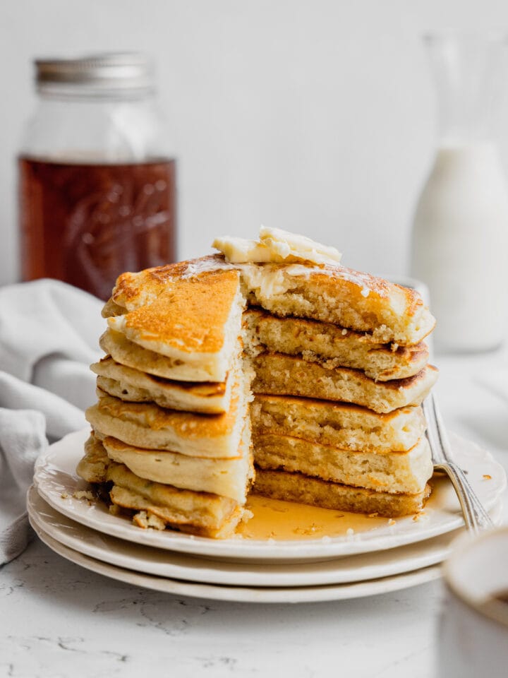 A stack of buttermilk pancakes on a white plate with a piece cut out to show the tender interior.