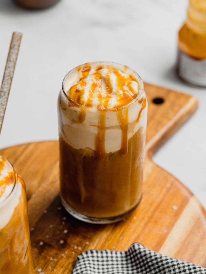 A glass filled with salted caramel cold foam cold brew coffee with a caramel drizzle sits on a wooden cutting board