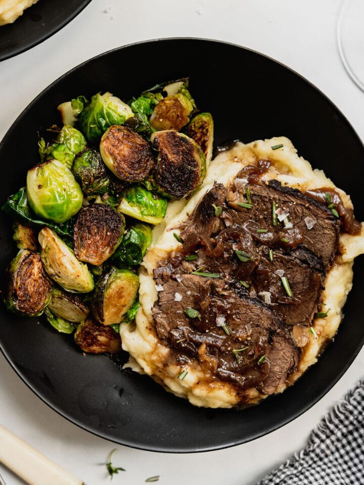 A black plate on a white background is filled with mashed potatoes, roasted vegetables, and topped with red wine venison roast.