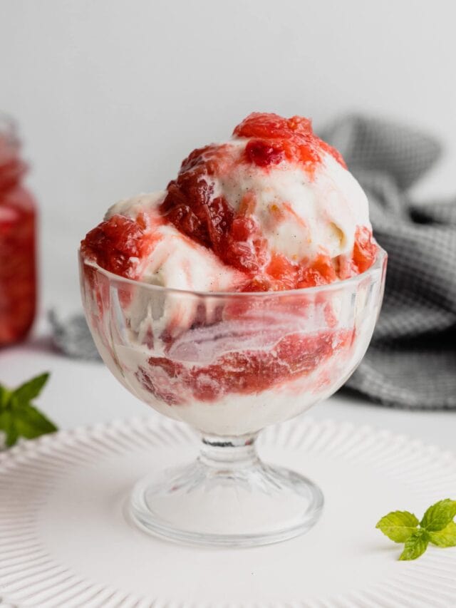 A dish of vanilla ice cream with rhubarb compote poured over the top. The dish sits on a white tray with a mint spring beside it.