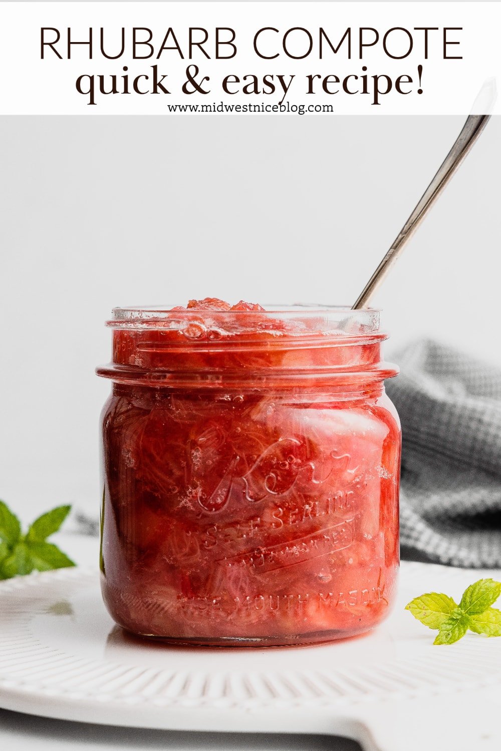 A glass jar filled with bright red rhubarb compote sits on a white tray with a spoon in the sauce. There is a mint sprig next to the jar.