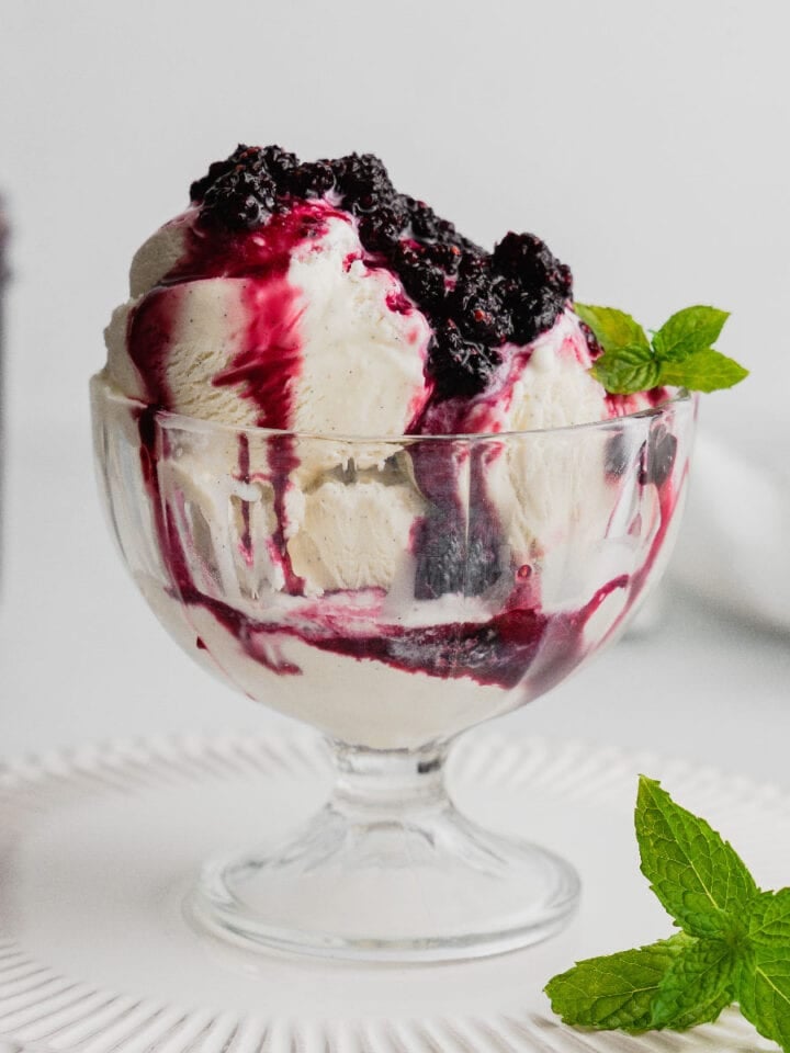 A glass dish filled with vanilla ice cream and topped with mulberry compote. There is a sprig of mint beside the dish and in the ice cream.
