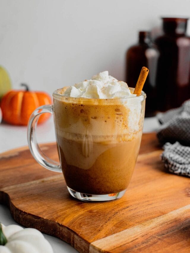 A mug filled with a homemade pumpkin spice latte and garnished with whipped cream and a cinnamon stick sits on cutting board.