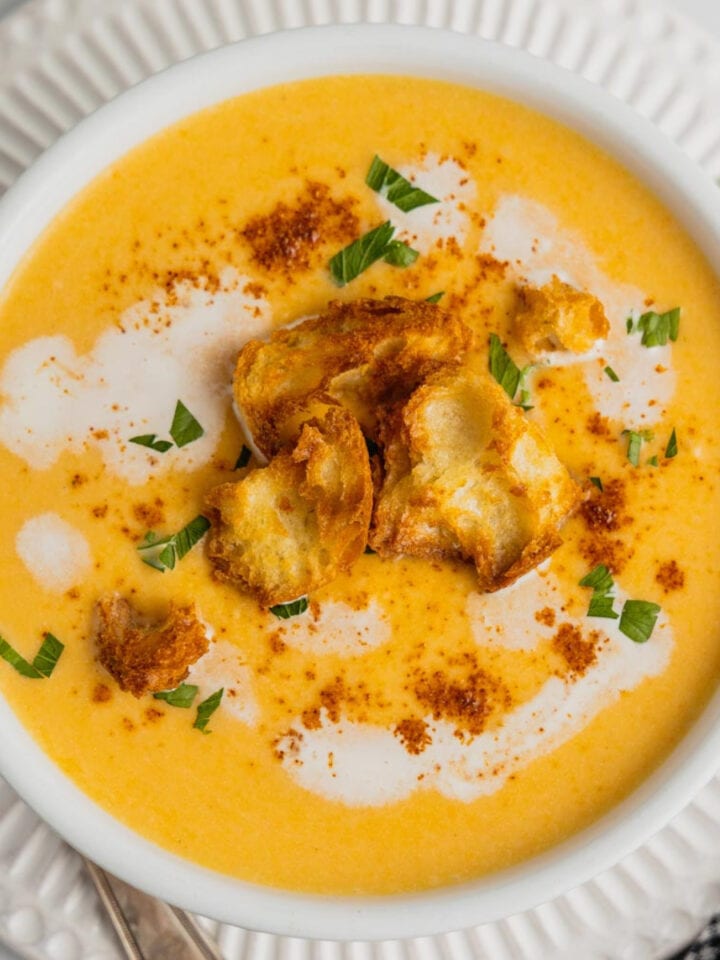 An overhead image of a white bowl filled with creamy acorn squash soup that is topped with croutons, parsley, and a drizzle of cream.