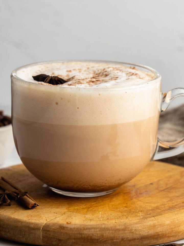 A classic chai latte in a glass mug on a round wooden board garnished with a star anise.