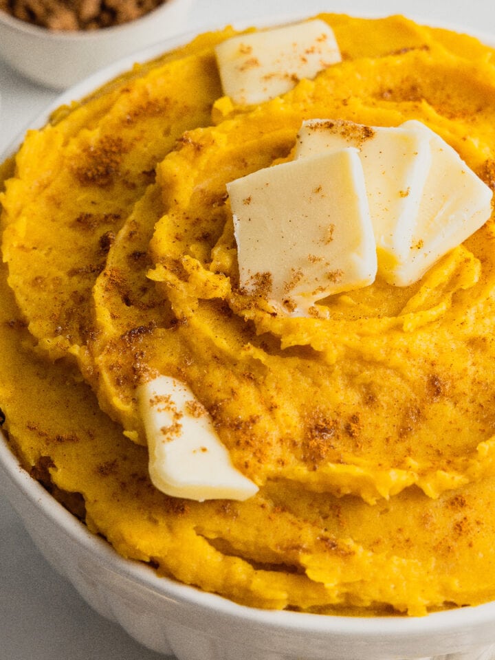 A white bowl filled with mashed acorn squash and topped with butter pats and a sprinkle of brown sugar and cinnamon.