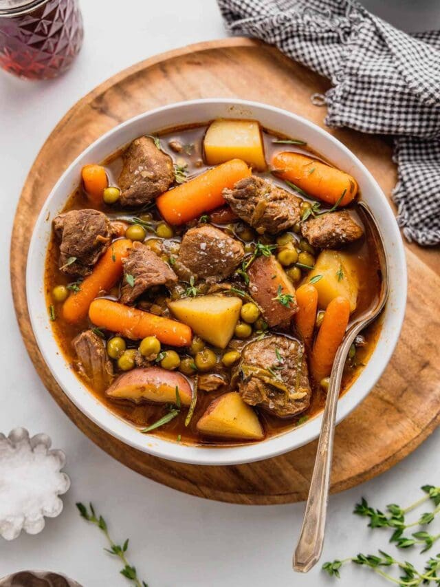 A bowl of easy venison stew sits on a round wooden board, there is a spoon in the bowl and checkered napkin beside it.