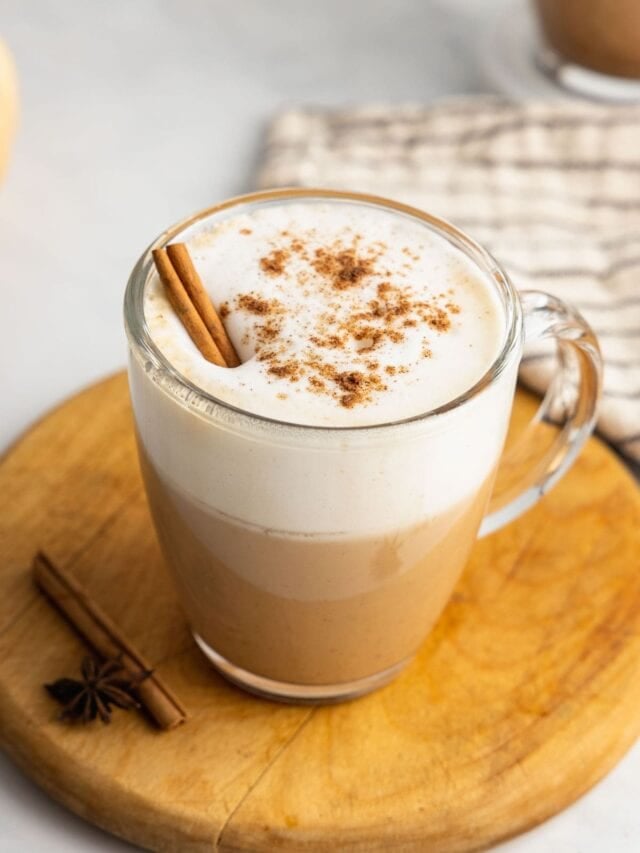 A pumpkin chai latte in a clear glass mug garnished with spices and a cinnamon stick sits on a round wooden board.
