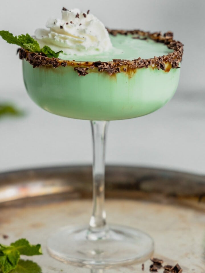 A green grasshopper cocktail sits in a coupe glass on a silver tray garnished with whipped cream and chocolate shavings.