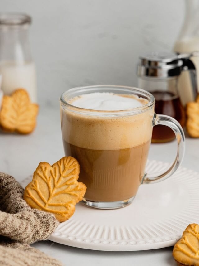 A maple leaf cookie rests against a clear glass mug filled with a maple latte.