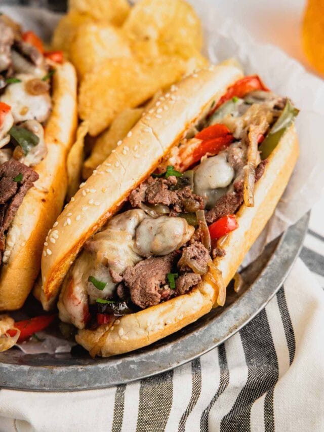 A venison cheesesteak sandwich sits on a metal tray with potato chips.
