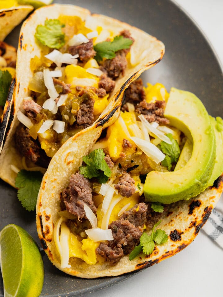 A close up of venison breakfast tacos on a gray plate topped with avocado and cilantro.