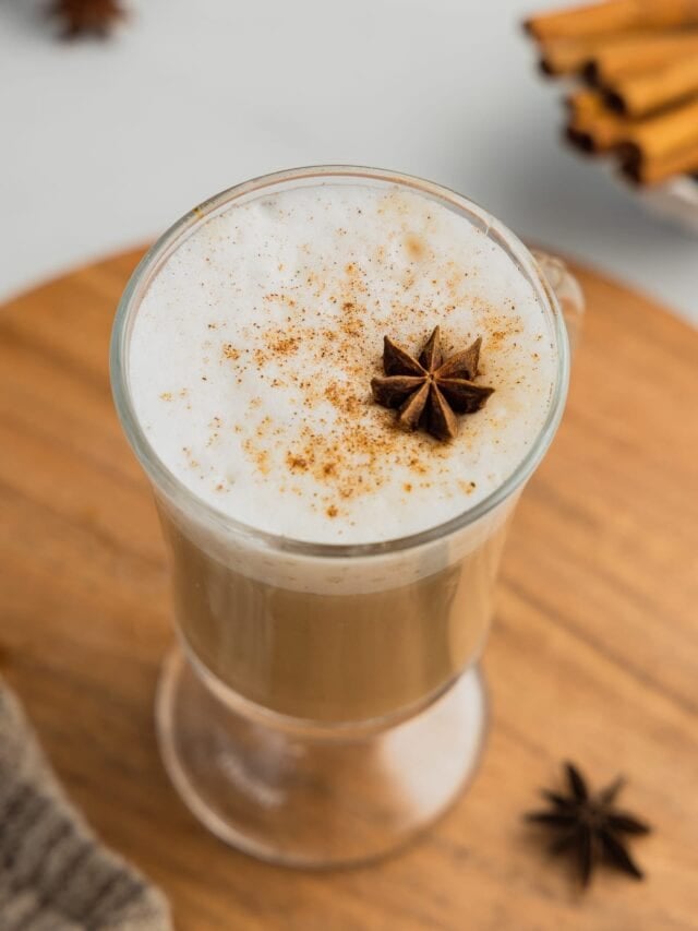 A clear mug filled with a vanilla chai latte garnished with chai spice and a star anise sits on a wooden board.