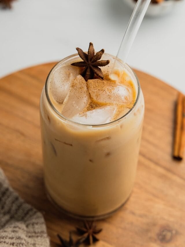 A glass filled with an iced vanilla chai latte garnished with a star anise and a clear straw.