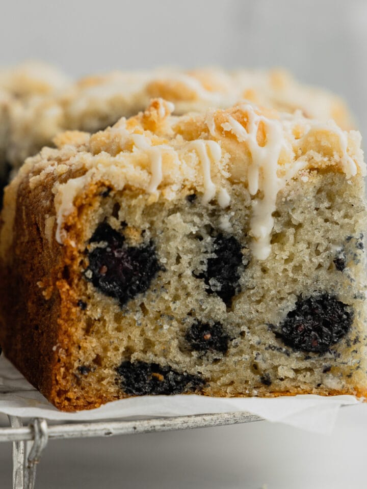 A slice of mulberry cake with crumb topping and vanilla glaze sits on a cooling rack.