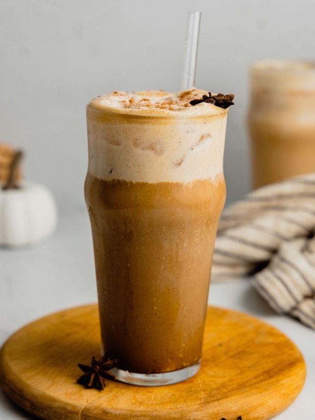 A glass of dirty chai latte topped with pumpkin cold foam sits on a wooden board.