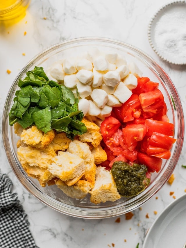 A glass bowl filled with tomatoes, basil, fresh mozzarella, and toasted bread plus more ingredients for a caprese panzanella salad.