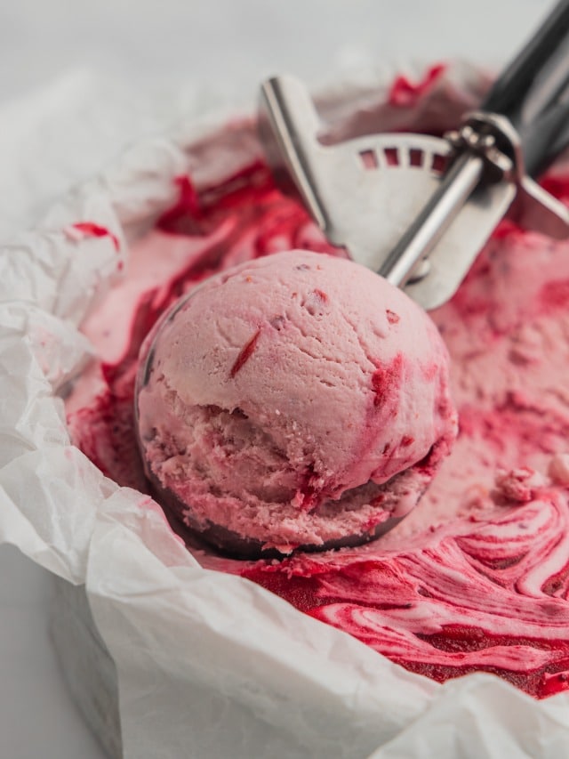 An ice cream scoop filled with cranberry ice cream in a cake pan filled with frozen no-churn ice cream.