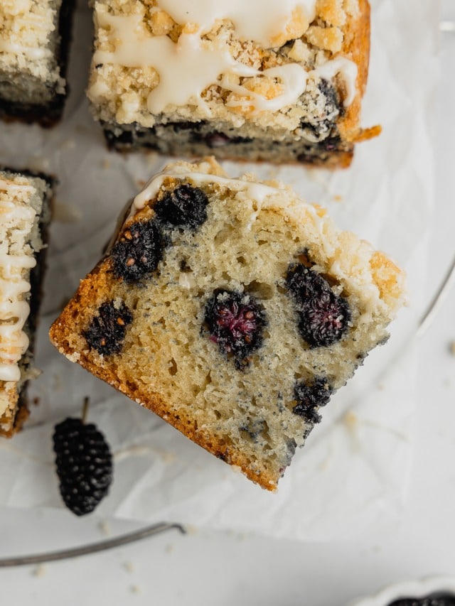 A slice of mulberry cake with crumb topping is set on its side next to the rest of the cake.