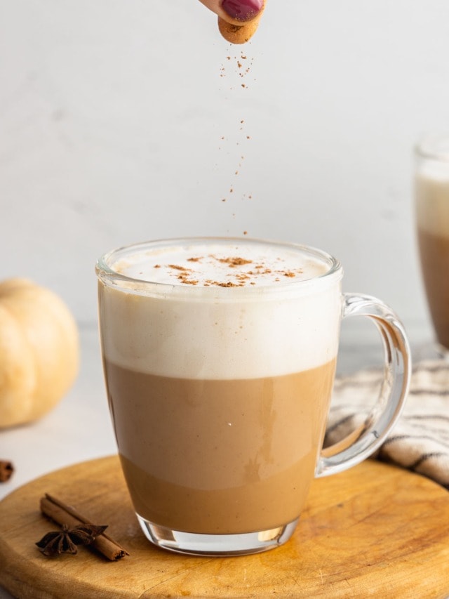 A hand sprinkles chai spice to garnish a pumpkin chai latte in a clear glass mug.