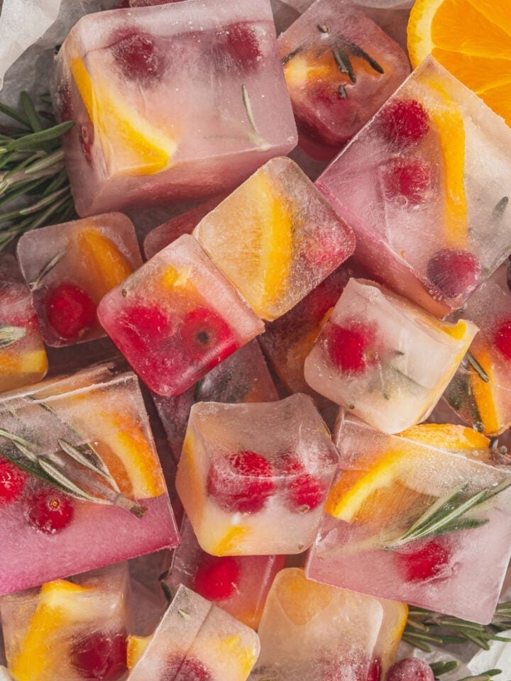 A pile of cranberry ice cubes sit on a parchment paper lined tray and are garnished with orange slices and fresh rosemary.
