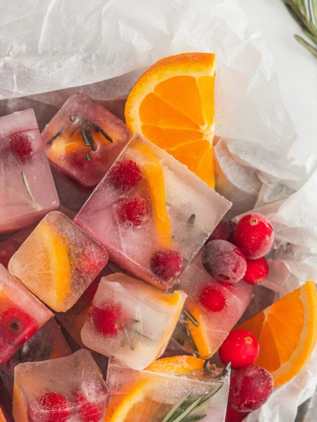 A pile of cranberry ice cubes on a parchment lined pan garnished with rosemary sprigs and orange slices.