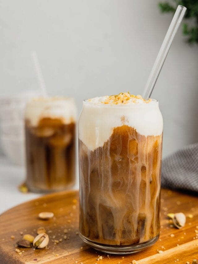 Two glasses of pistachio cream cold foam cold brew are show, one on a wooden board in the foreground, one in the background.