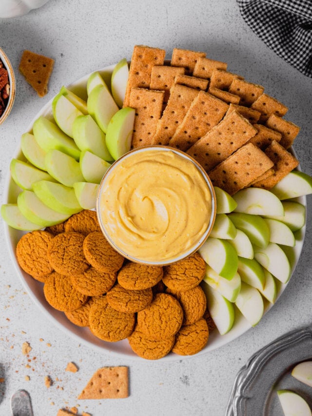 A small bowl of pumpkin pie cheesecake dip sits surrounded by cookies and cut apples for dipping.