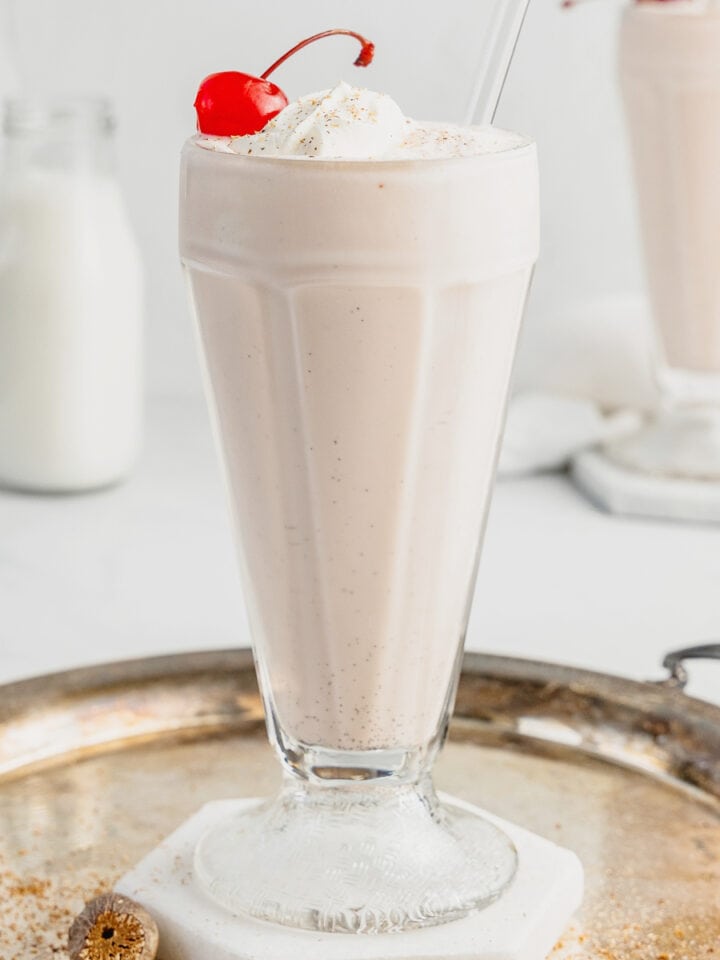 A glass filled with a pink squirrel made with ice cream and garnished with a maraschino cherry sits on a coaster on a silver tray.