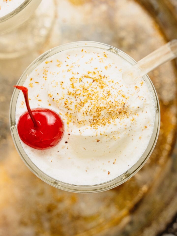 An overhead image an ice cream brandy alexanders with a cherry on top and a straw. The glass is resting on a silver tray.