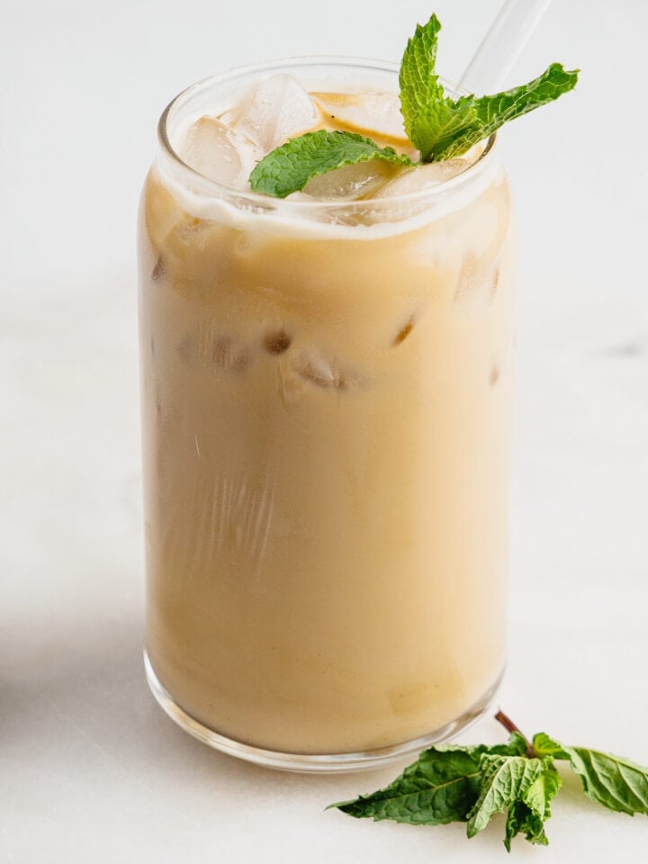 A glass filled with peppermint iced latte that is garnished with three mint leaves on top. There is a clear straw in the glass and three mint leaves displayed beside the glass.