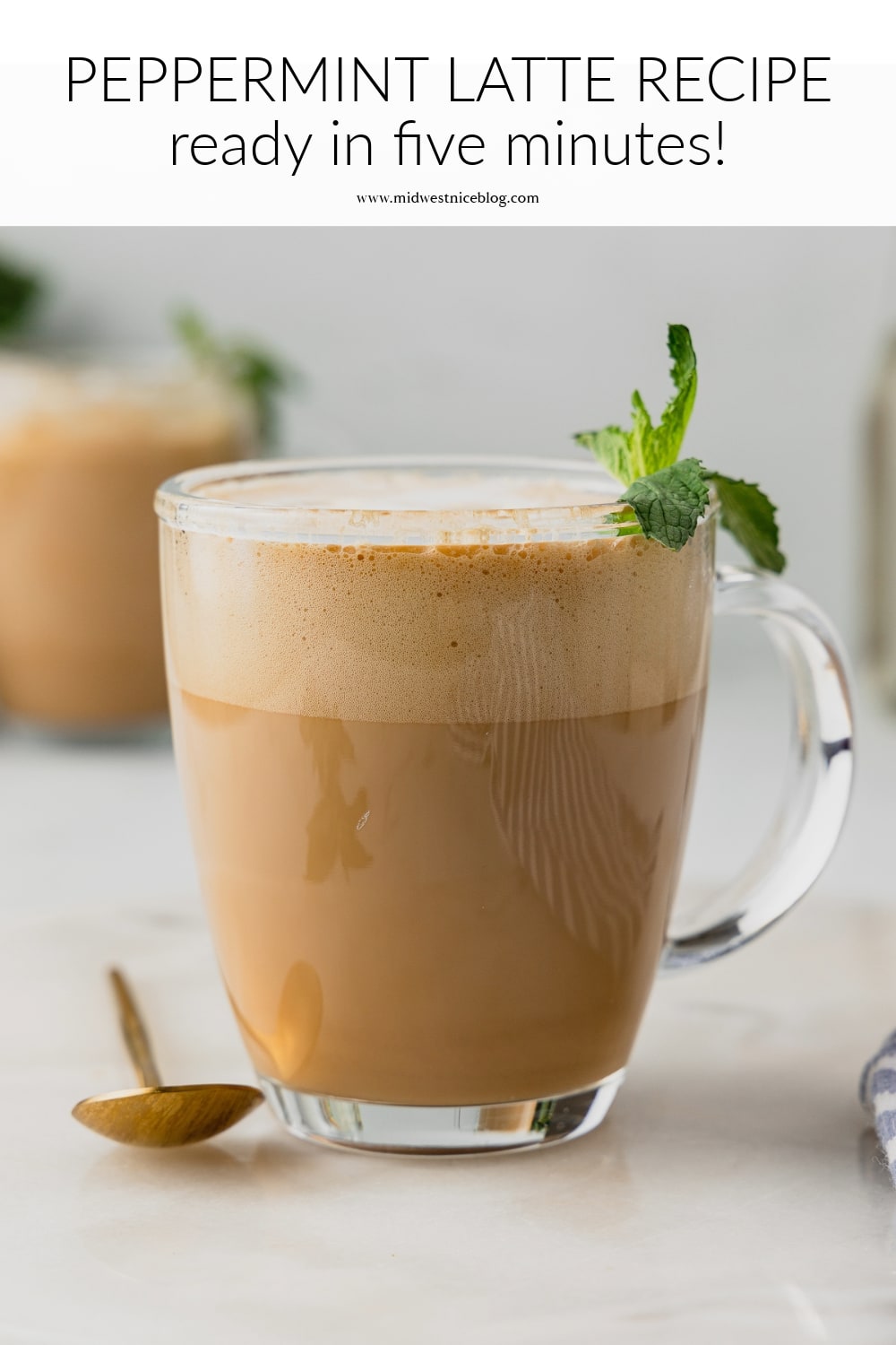 A peppermint latte in a glass mug garnished with mint leaves on top. There's a golden spoon next to the mug and a second peppermint latte in the background slightly out of focus.