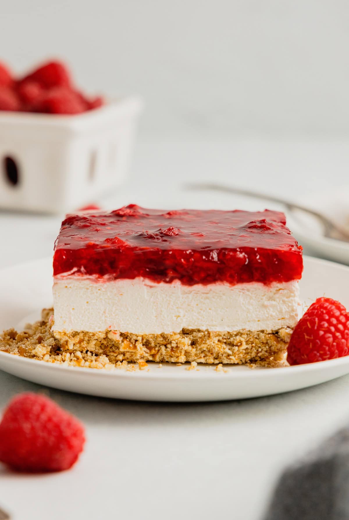 A slice of raspberry pretzel jello salad sits on a white plate garnished with fresh raspberries.