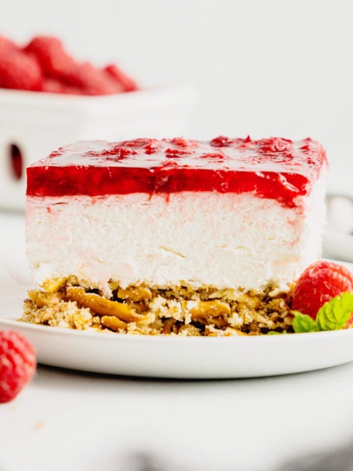 A slice of raspberry pretzel jello salad sits on a white plate garnished with fresh raspberries.
