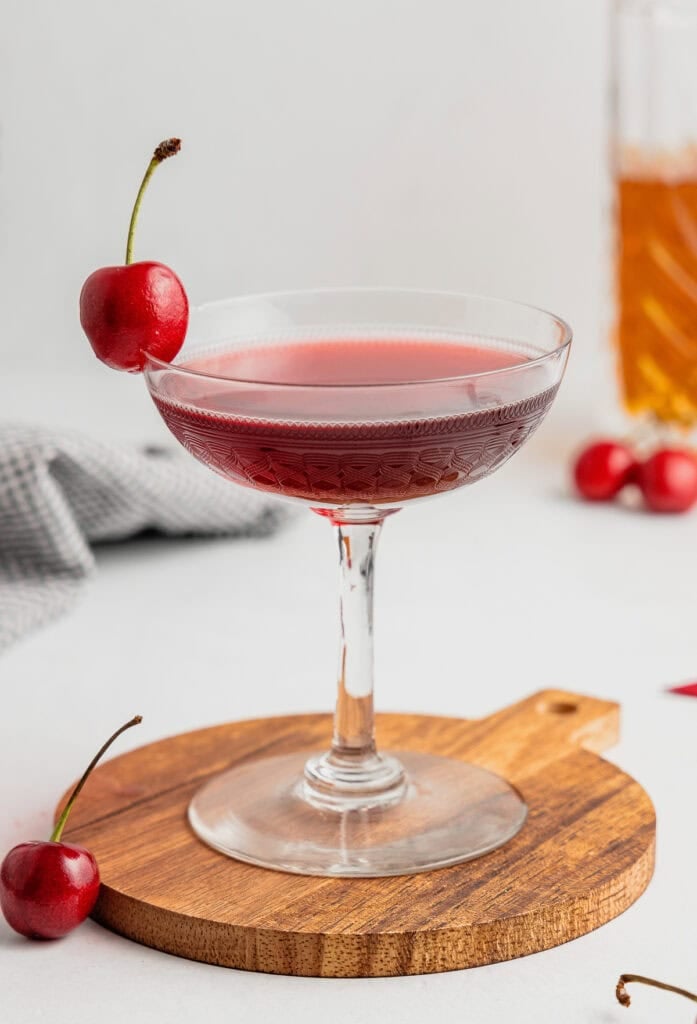 A coupe glass filled with cherry bounce and garnished with a fresh cherry set on a wooden coaster.