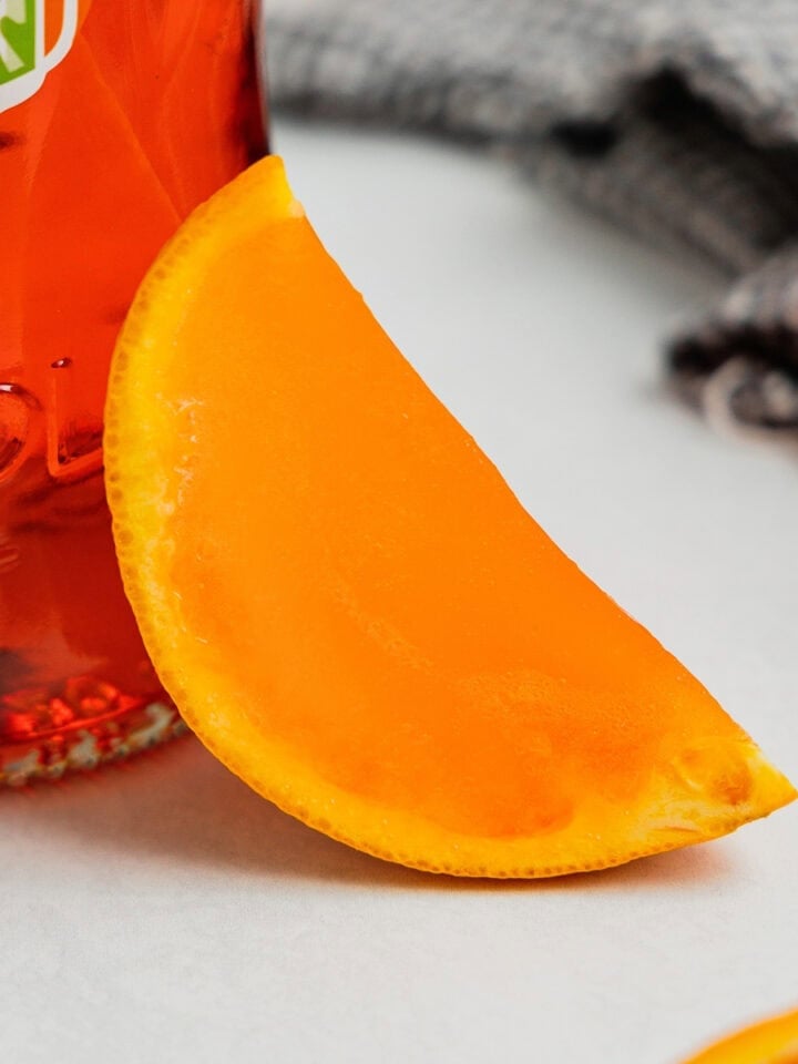 An aperol spritz jello shot cut into an orange wedge rests against a bottle of the Italian apertif.