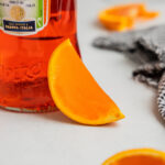An aperol spritz jello shot cut into an orange wedge rests against a bottle of the Italian apertif.