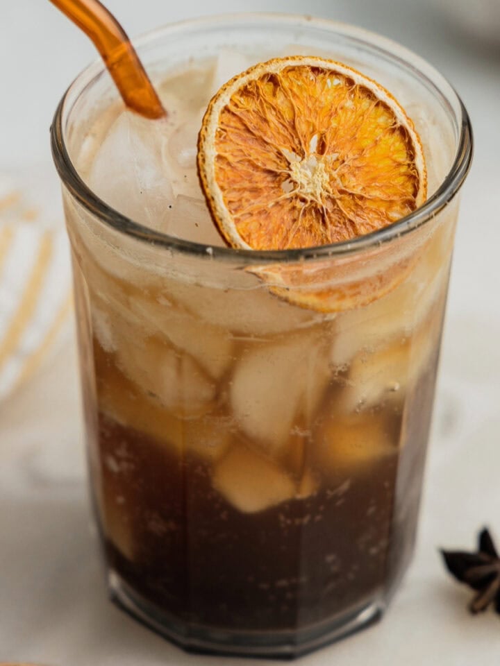 A glass filled with coffee soda, ice, an orange slice, and a glass orange straw.